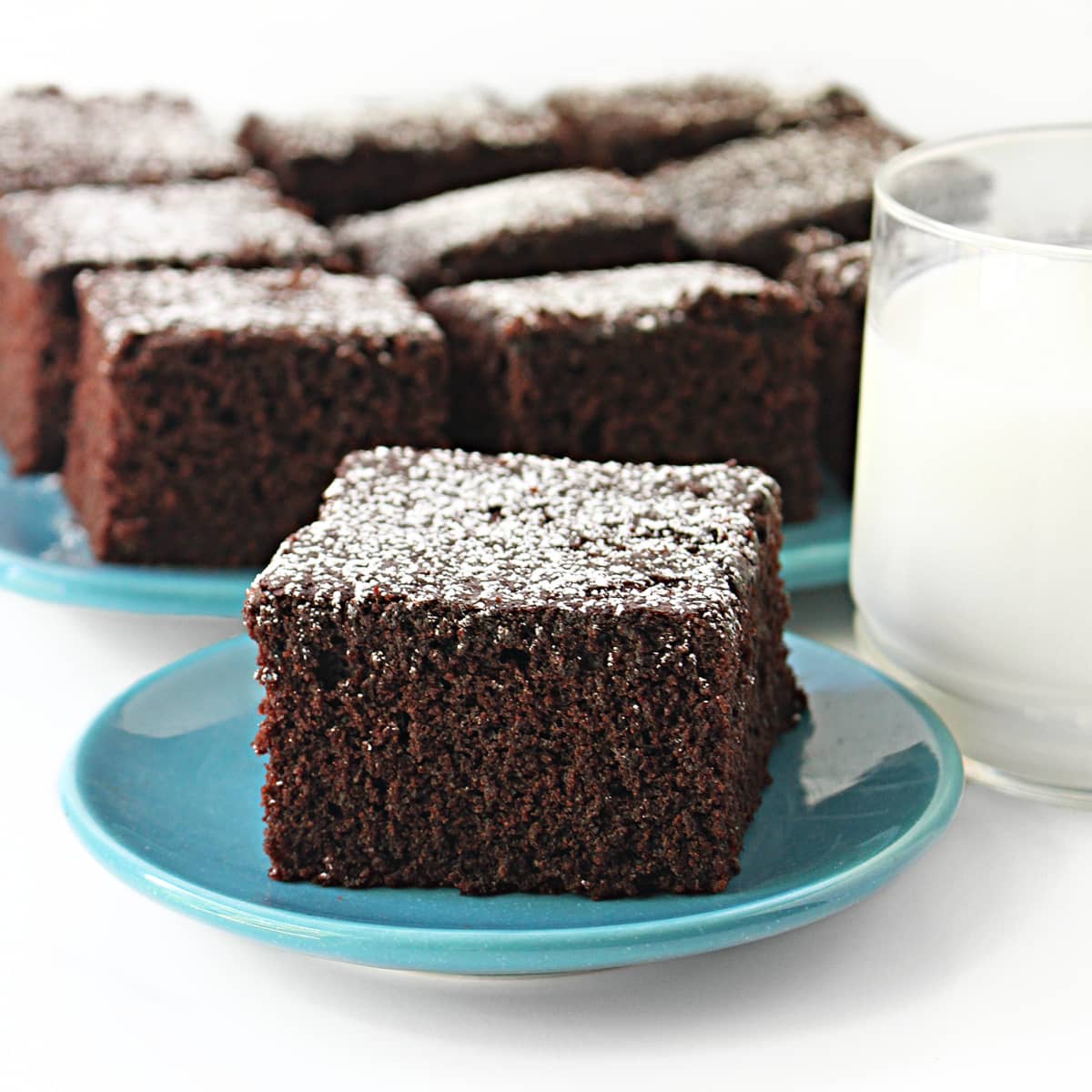 A slice of Chocolate Snack Cake topped with powdered sugar on a plate with a glass of milk.
