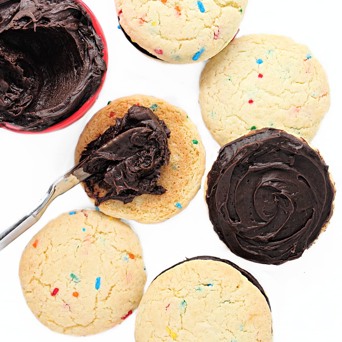 Chocolate frosting being spread on the bottom of a chewy Funfetti Cake Mix Cookie to make sandwich cookies.